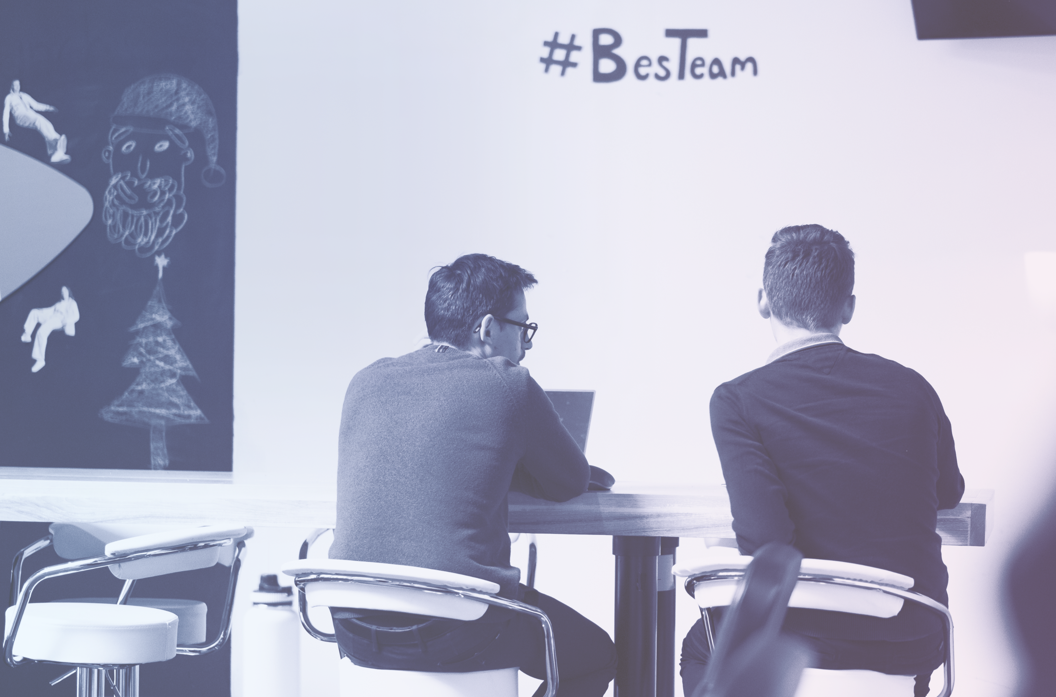 Two people sitting at a table with a whiteboard in the background displaying '#Besteam' and some drawings.